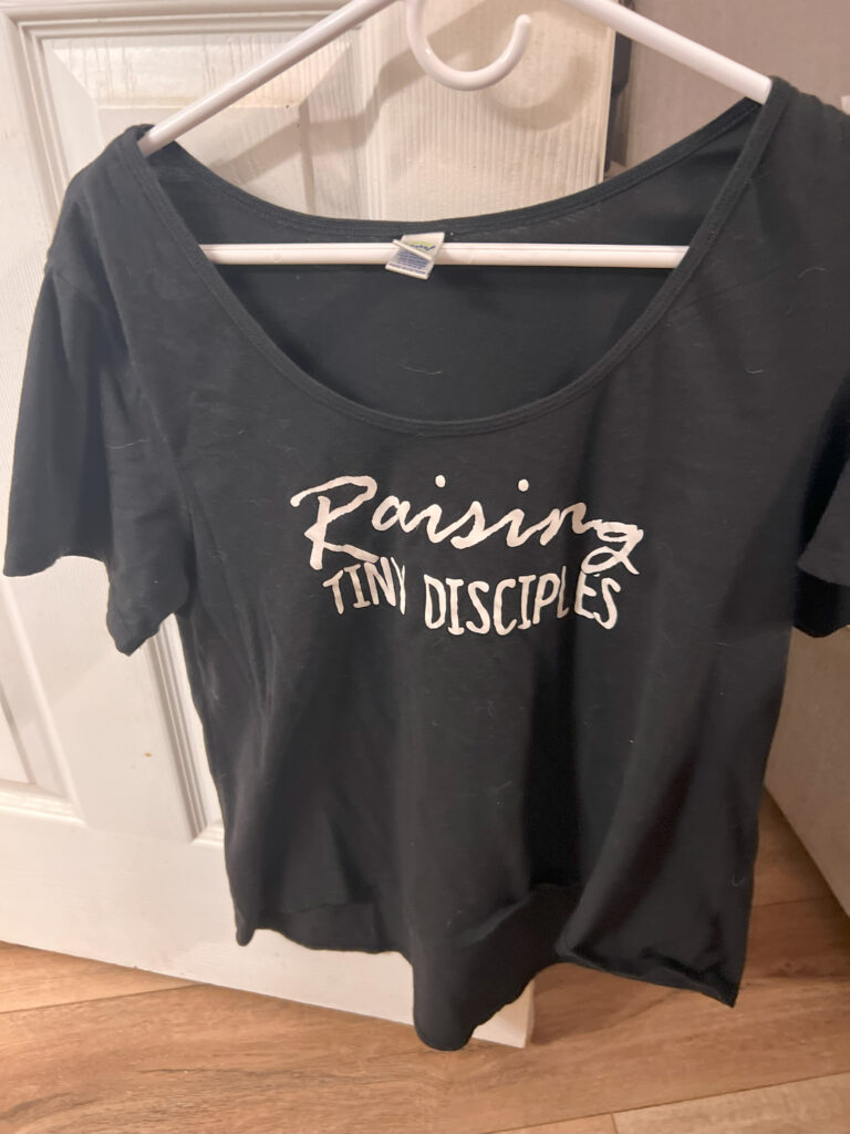 Black T-shirt with white “Raising Tiny Disciples” lettering hanging on a hanger, representing a homemade HTV mom shirt from 2016.